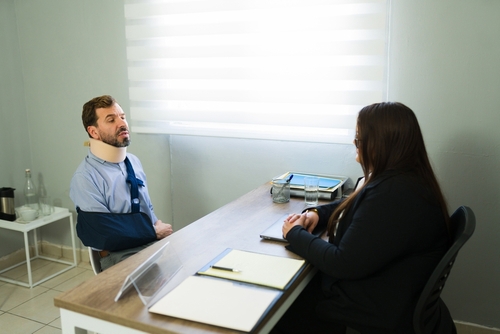 Man wearing neck brace and arm sling, grimacing beside hospital bed